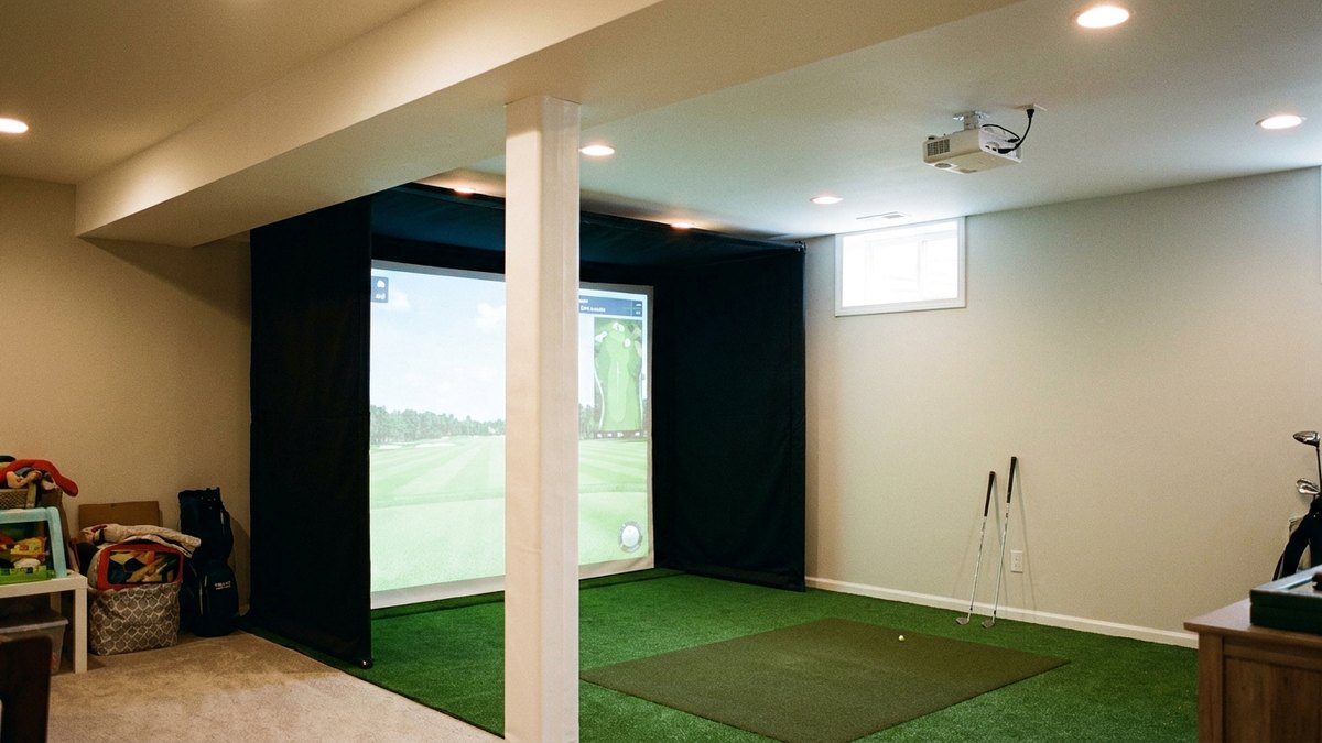 Concept render of a basement golf simulator with wall-to-wall turf, a matte black screen wall, and a ceiling-mounted projector.
