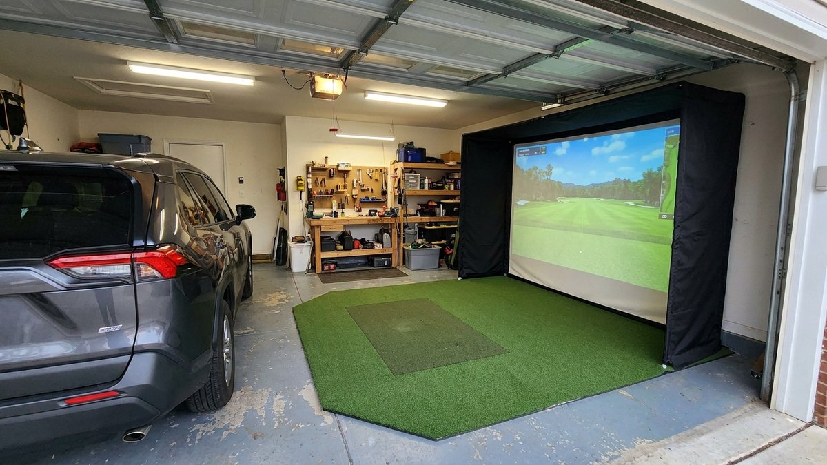 Concept render of a golf simulator built into one bay of a two-car garage, with a corner turf pad and a matte black backdrop panel.
