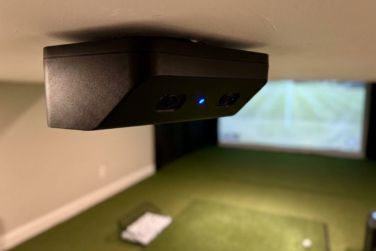 Detail view of an overhead launch monitor mounted above a hitting mat with ball data displayed on a nearby monitor.